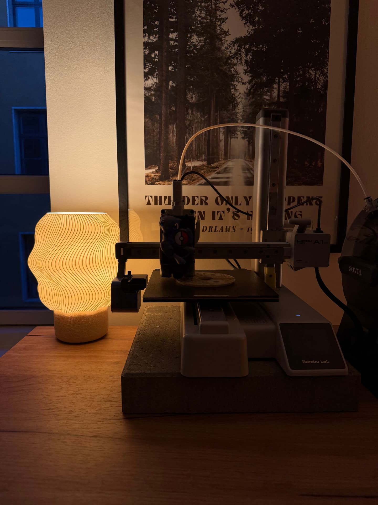 Tændt riflet SETTLE-lampe ved siden af en 3D-printer der printer en lampedel om aftenen